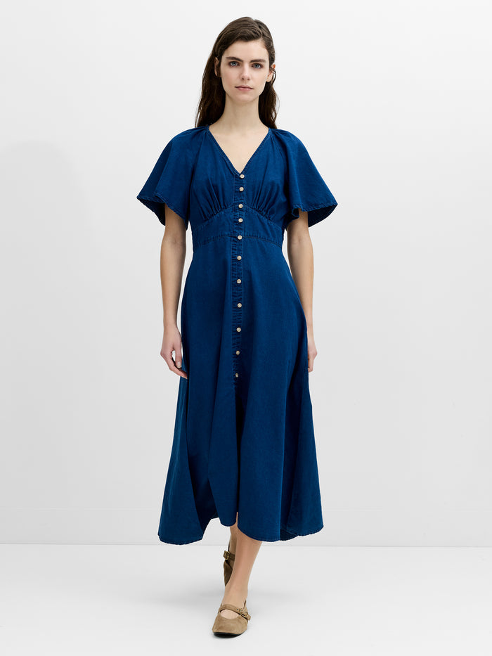 A woman wearing a flowing, midi length navy blue dress with a V-neck, flutter sleeves and an empire waistline with button down fastening.